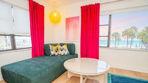 Une pièce avec un canapé vert, une table ronde en verre, des rideaux rouges, un luminaire jaune et une vue sur la plage.