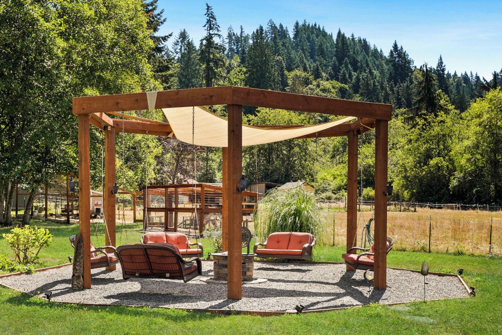A wooden gazebo with a swing set and seating area, surrounded by a grassy field and trees.