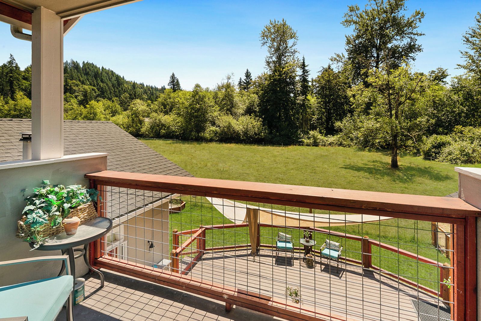 A balcony with a view of a green lawn and trees, a table with chairs, and a potted plant.