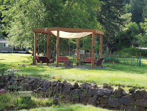 A wooden gazebo with a canopy, surrounded by a stone wall, in a grassy backyard with trees and a house in the background.