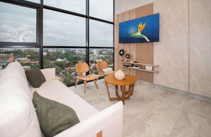 Ein modernes Wohnzimmer mit großem Fenster, einer Couch, zwei Sesseln, einem runden Tisch und einem wandmontierten Fernseher.