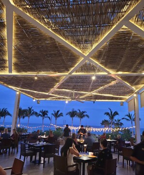 An outdoor dining area with tables and chairs, a thatched ceiling, and palm trees in the background. - Villa La Valencia Cabo Luxury 2Bedroom 3Bathroom Suite with Stunning Ocean Views (San José del Cabo)