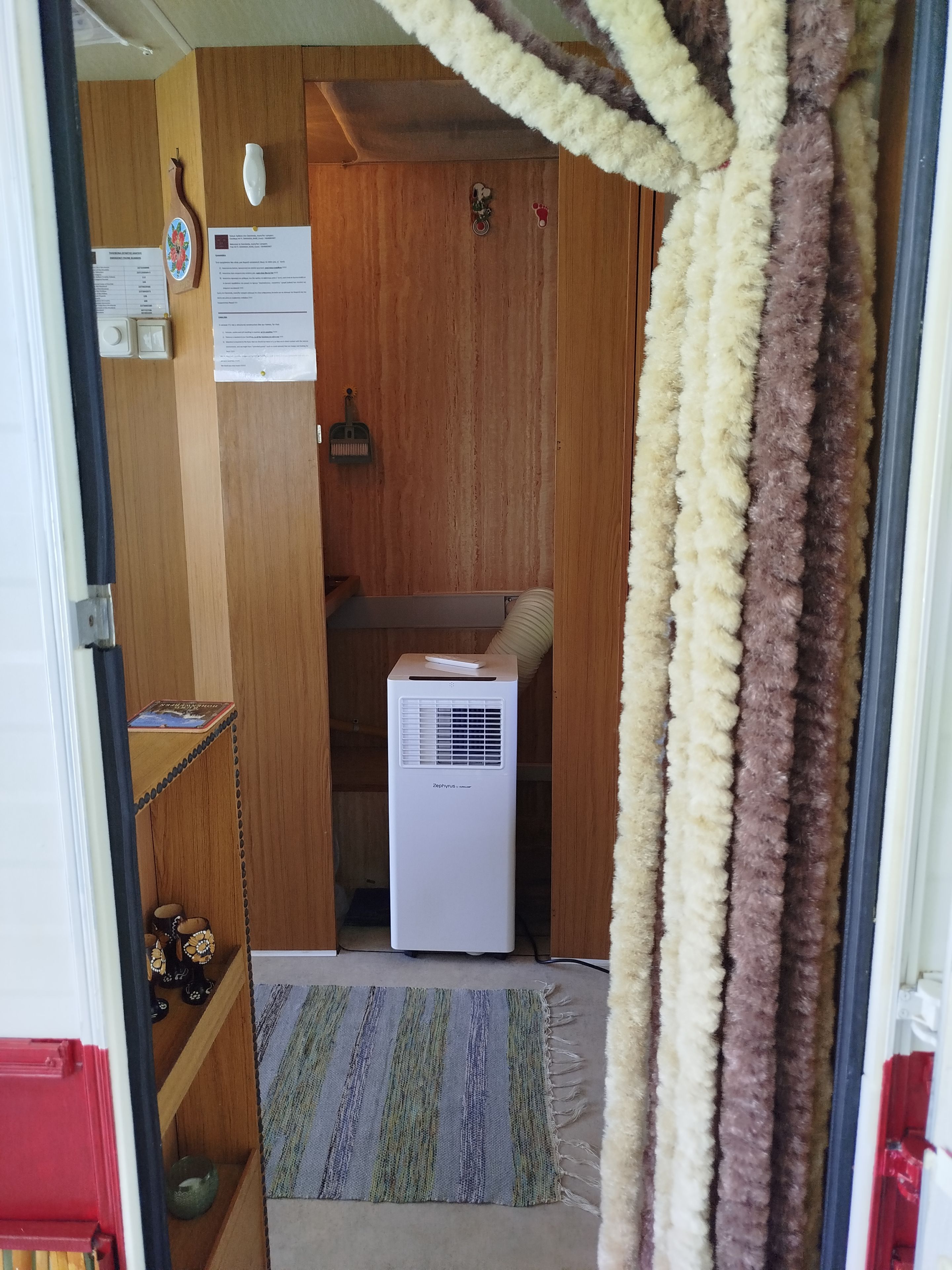 Una habitación con puerta de madera, aire acondicionado blanco, alfombra a rayas y una estantería con adornos.