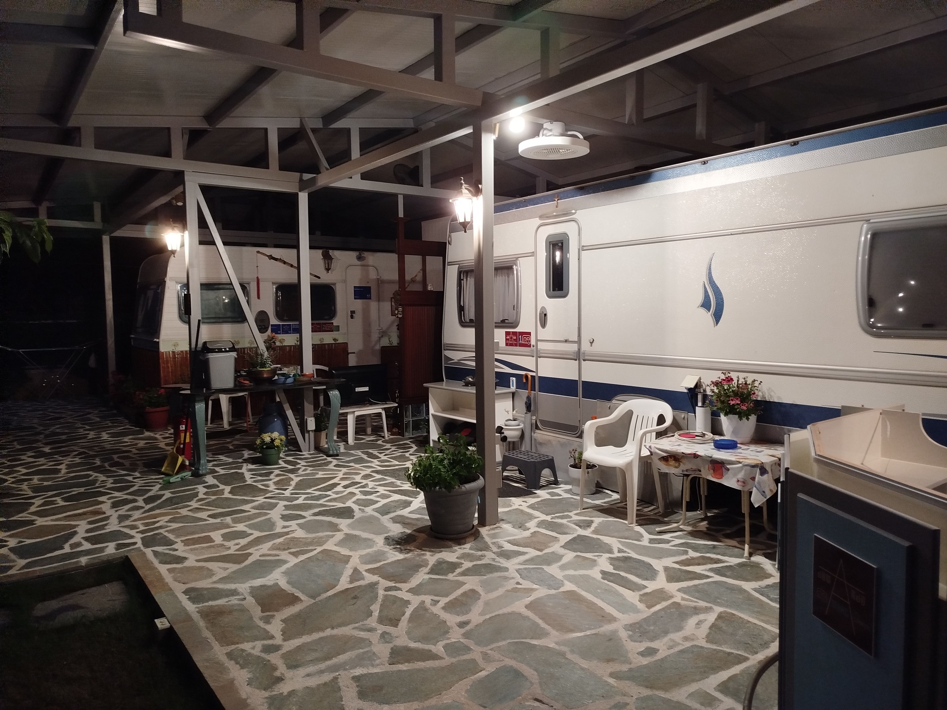 Un espace extérieur abrité comprenant une table à manger, des chaises, des plantes en pot et un camping-car blanc avec une bande bleue.