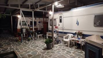 Un espace extérieur abrité comprenant une table à manger, des chaises, des plantes en pot et un camping-car blanc avec une bande bleue.