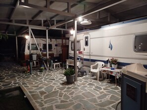 A covered outdoor area with a dining table, chairs, potted plants, and a white RV with a blue stripe.
