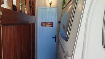 Un couloir étroit avec une porte bleue portant l’inscription « Toilettes de M. Ávila », une rambarde en bois sur la gauche et un camping-car blanc garé sur la droite.