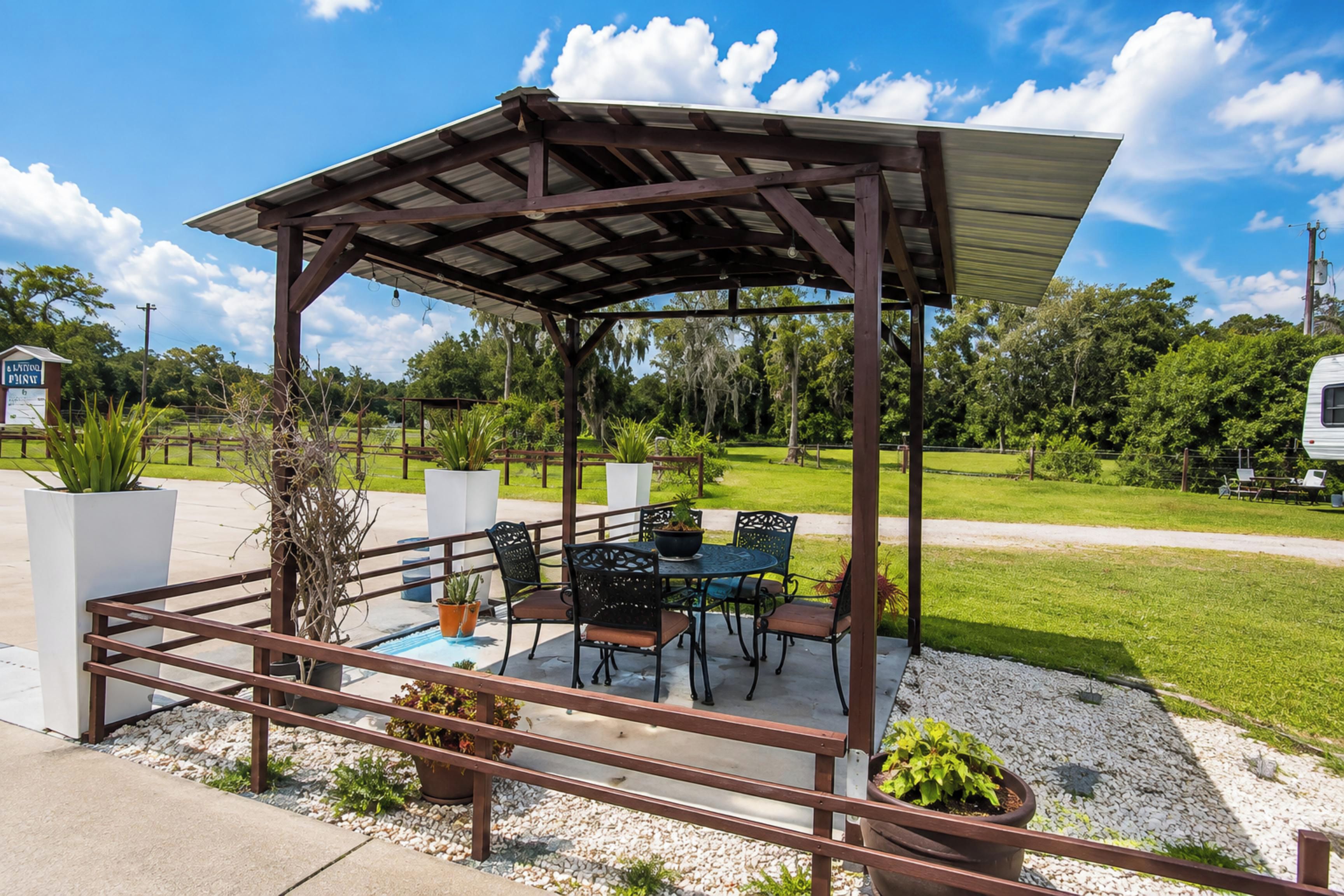 A covered outdoor seating area with tables and chairs, surrounded by greenery and a clear sky.