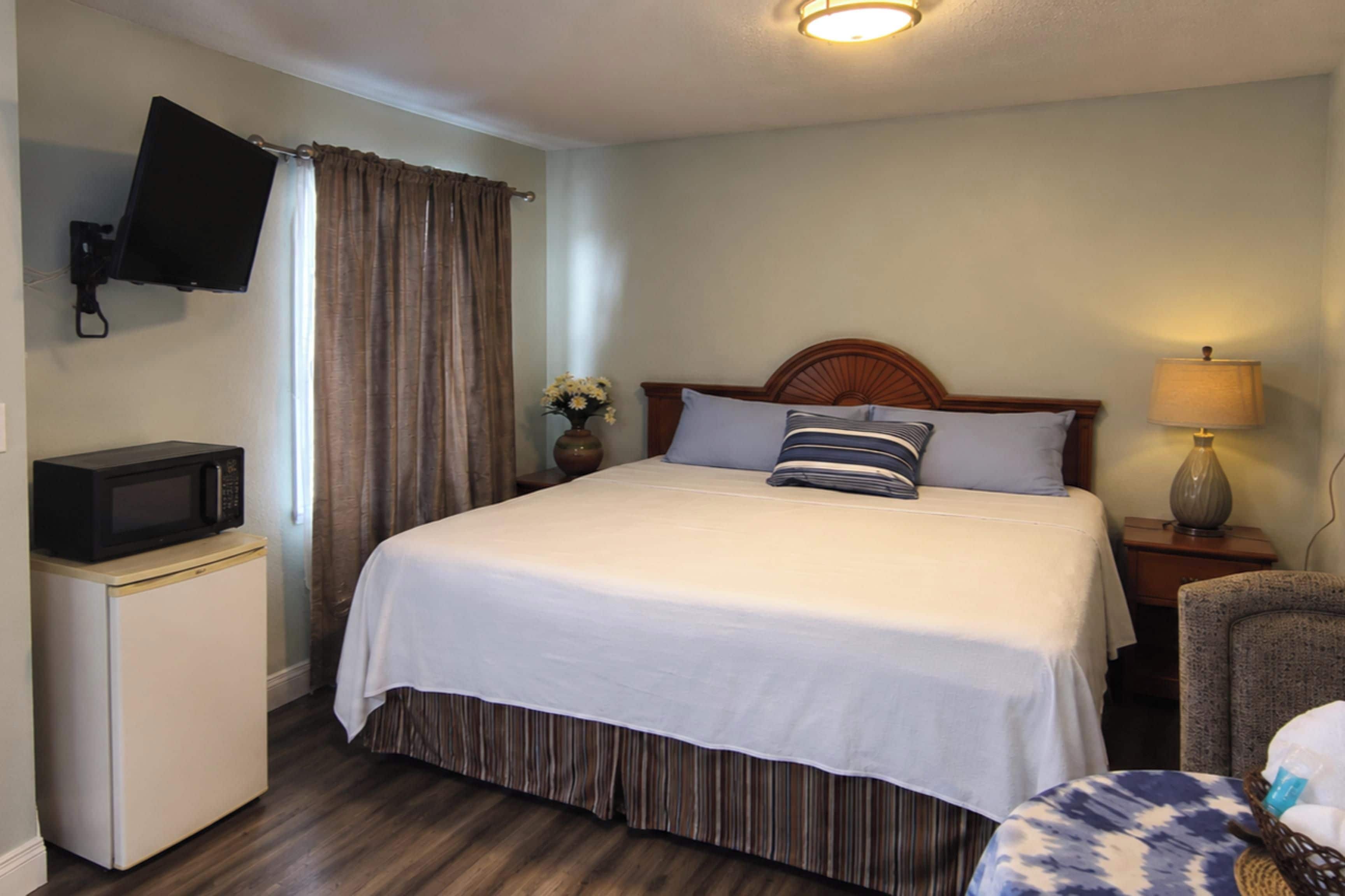 Quarto de hotel com cama, televisão na parede, secretária pequena com abajur e uma cadeira.