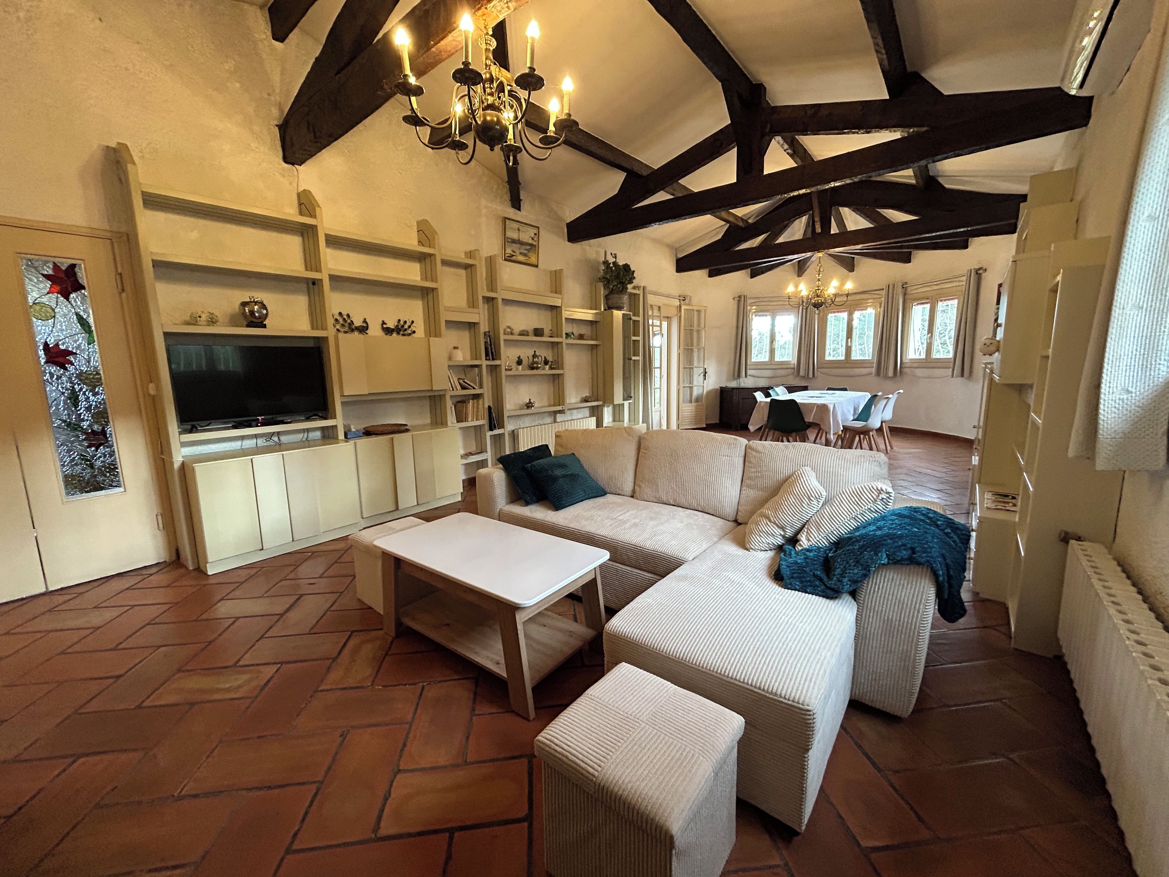 A spacious living room with a large sofa, a television, and a fireplace. There are wooden beams on the ceiling and a chandelier.