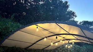 Et udendørs spiseområde med et bord og stole under en stor parasol, omgivet af grønt.