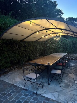 Ein Essbereich im Freien mit Tisch und Stühlen unter einem großen Sonnenschirm, umgeben von viel Grün.