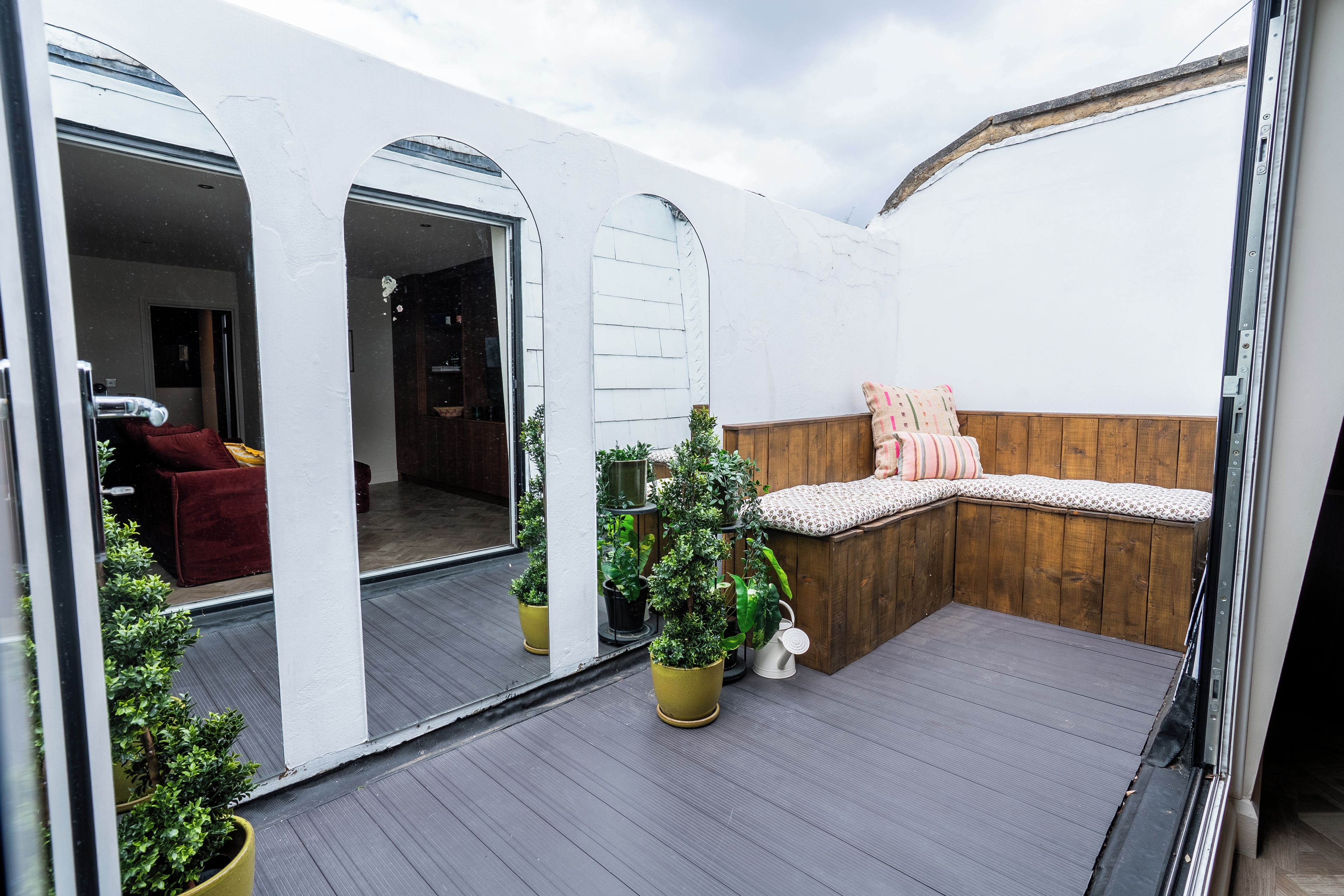 A covered outdoor seating area with a wooden bench and cushions, surrounded by potted plants.