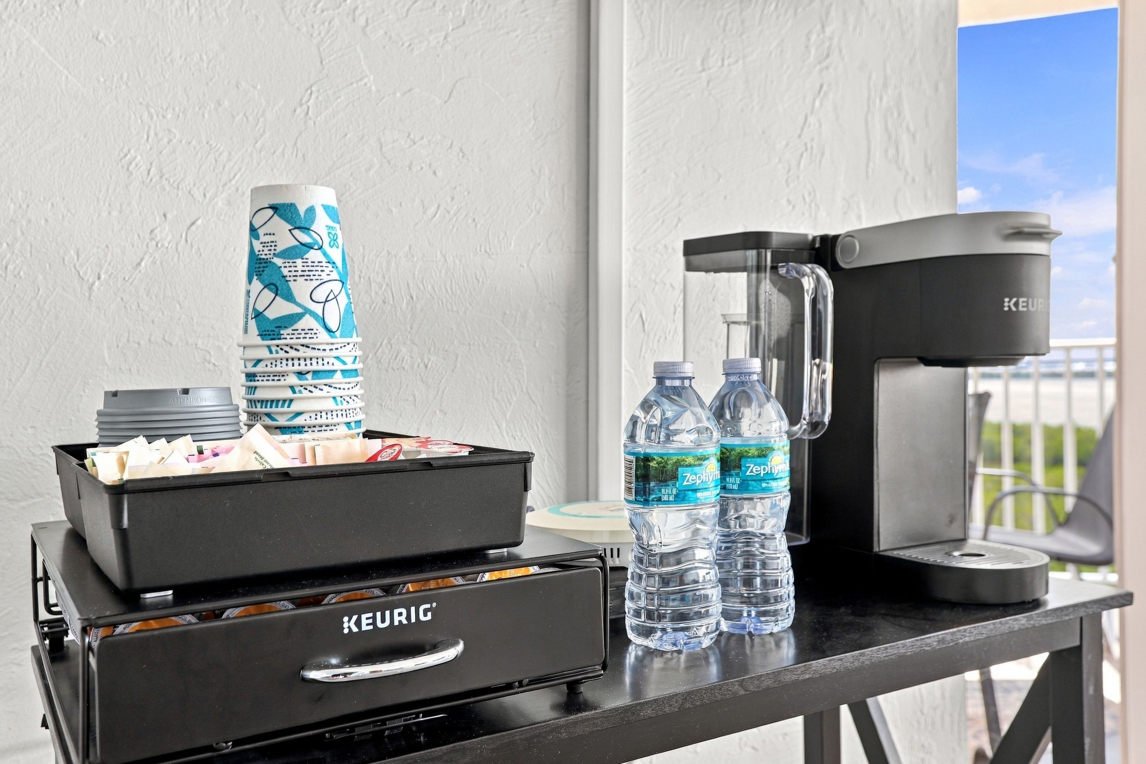 Una cafetera Keurig, dos botellas de agua, una pila de vasos y un cubo de basura sobre una mesa.