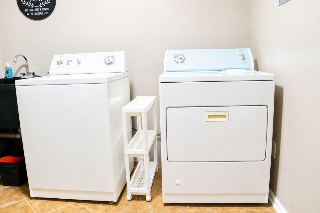 A laundry room with two white washing machines, a small white shelf, and a black trash bin.
