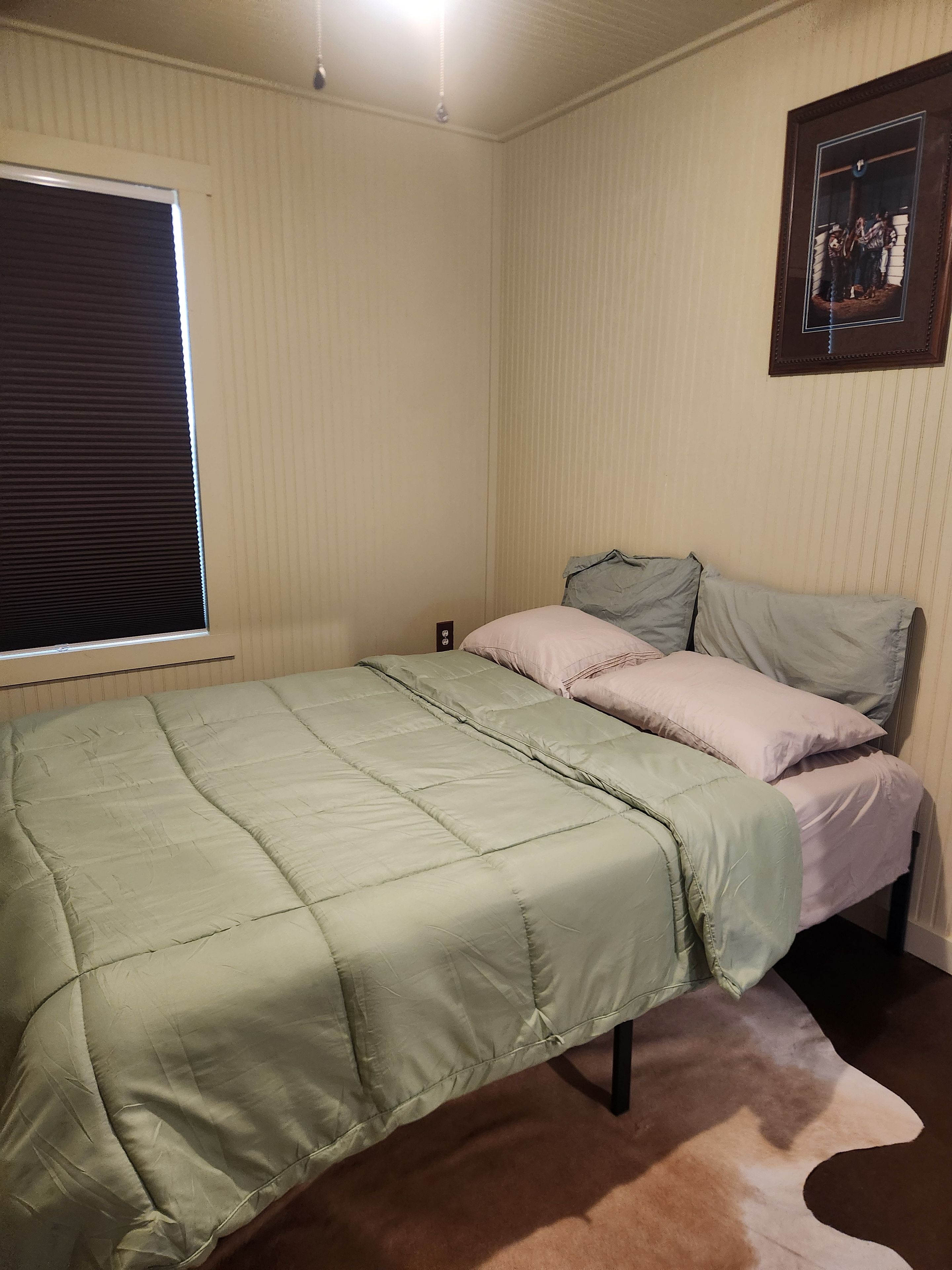 Uma cama de solteiro com colcha verde e duas almofadas, uma janela com persianas e um quadro na parede.