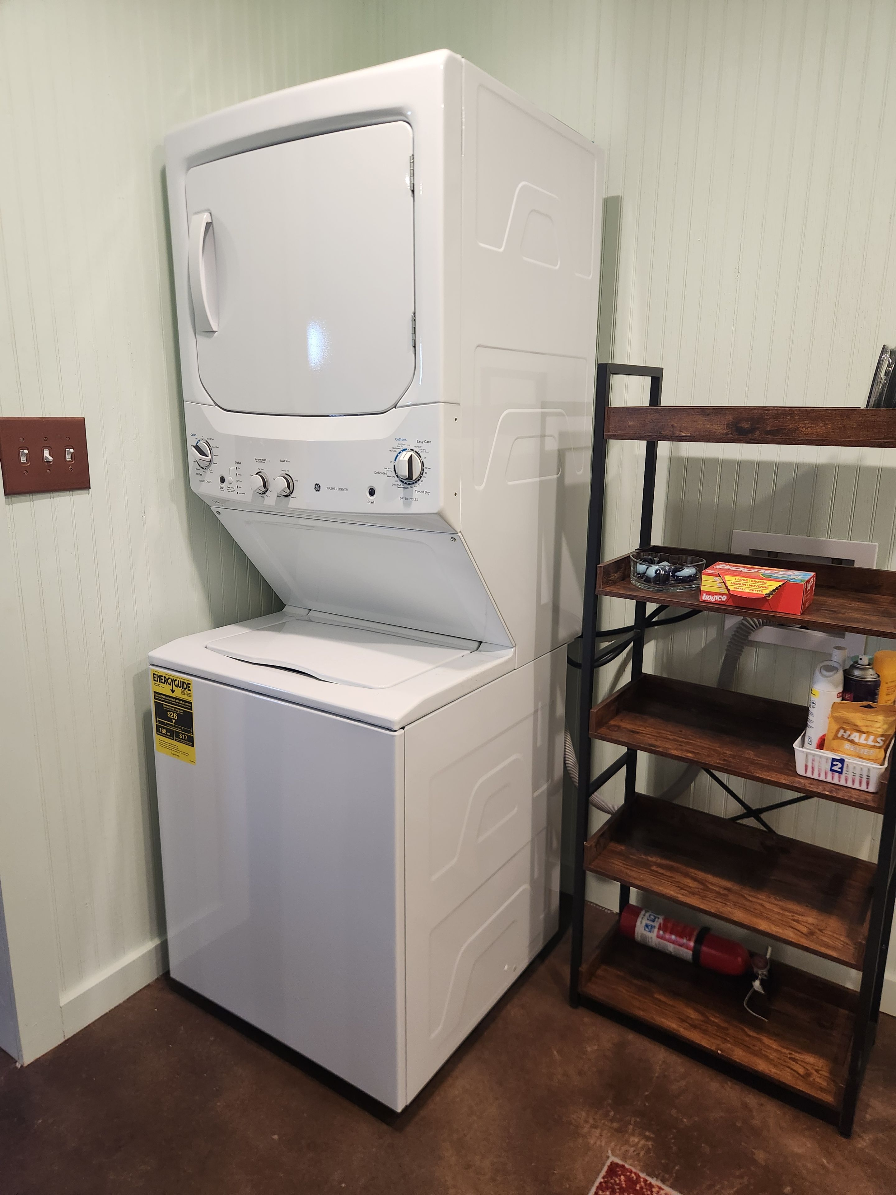 Uma máquina de lavar e secar compacta com painel de controle, posicionada ao lado de uma prateleira de madeira com diversos objetos.