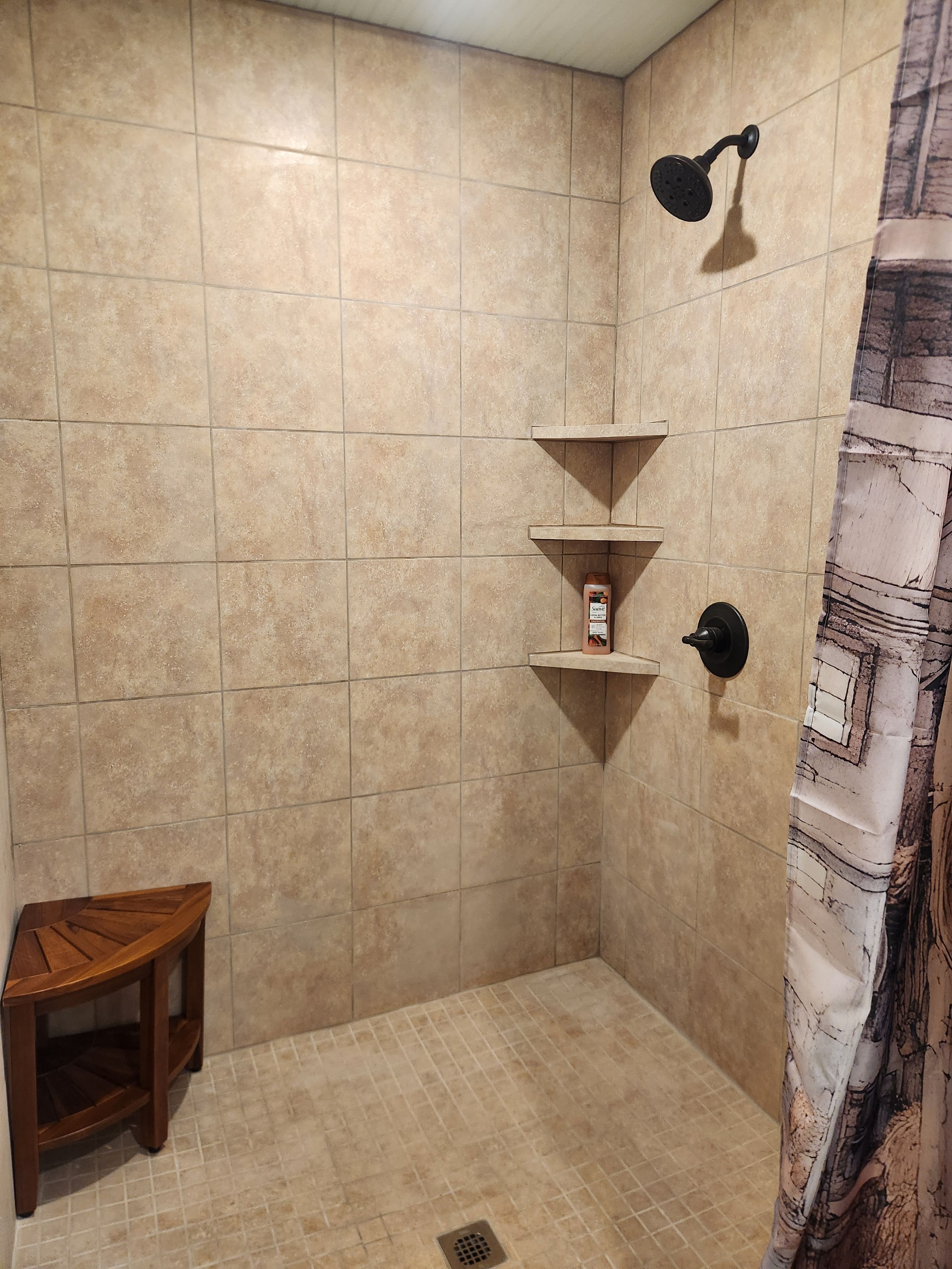 Zona de ducha con baldosas color beige, un taburete de madera y un cabezal de ducha.