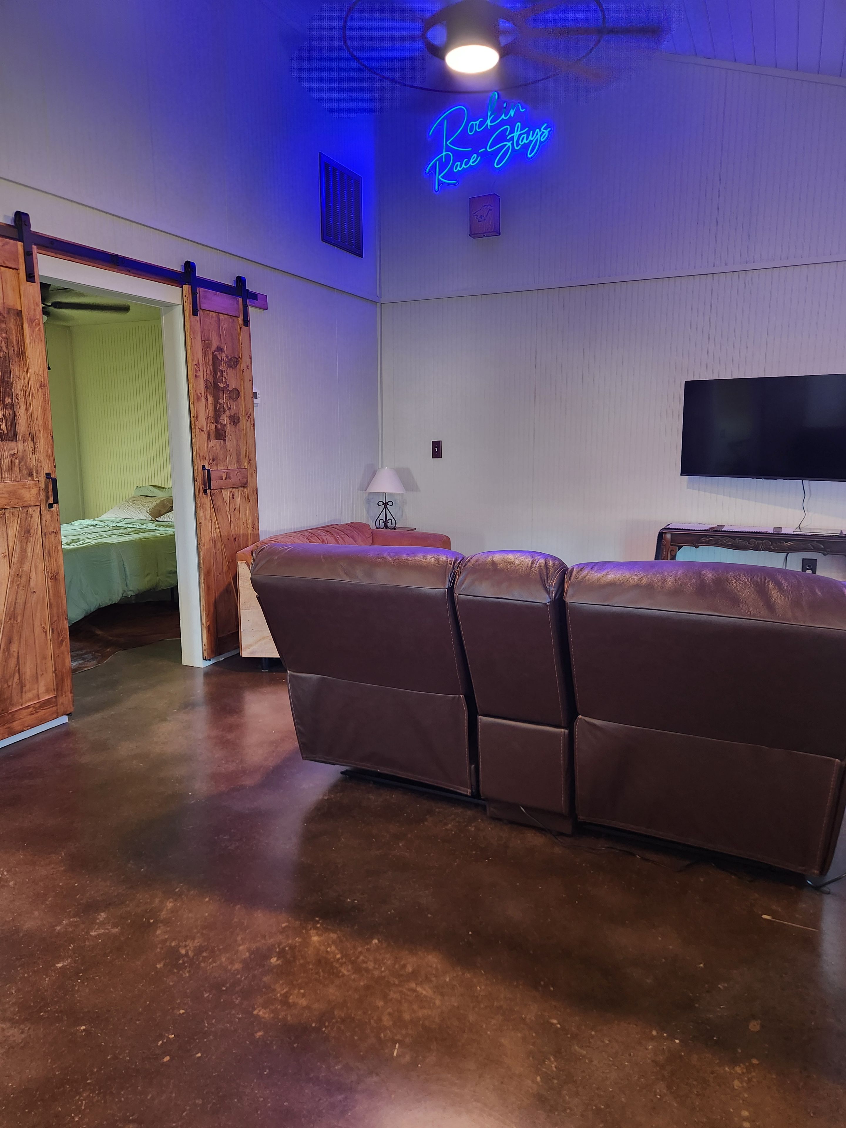 A room with a large brown leather sofa, a sliding barn door, a flat-screen TV, and a neon sign reading 'Rockin' Race-Stays'.