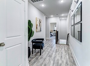 A hallway with a wooden floor, a black chair, a potted plant, and a painting on the wall. - Luxury Modern Stay Minutes from NRG & Med Center (Houston)