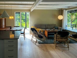 Ein geräumiger Wohnbereich mit Esstisch, Sofa und Sesseln, ein großes Fenster mit Blick auf Grünflächen und eine Küchenzeile mit Schränken und Arbeitsplatte.