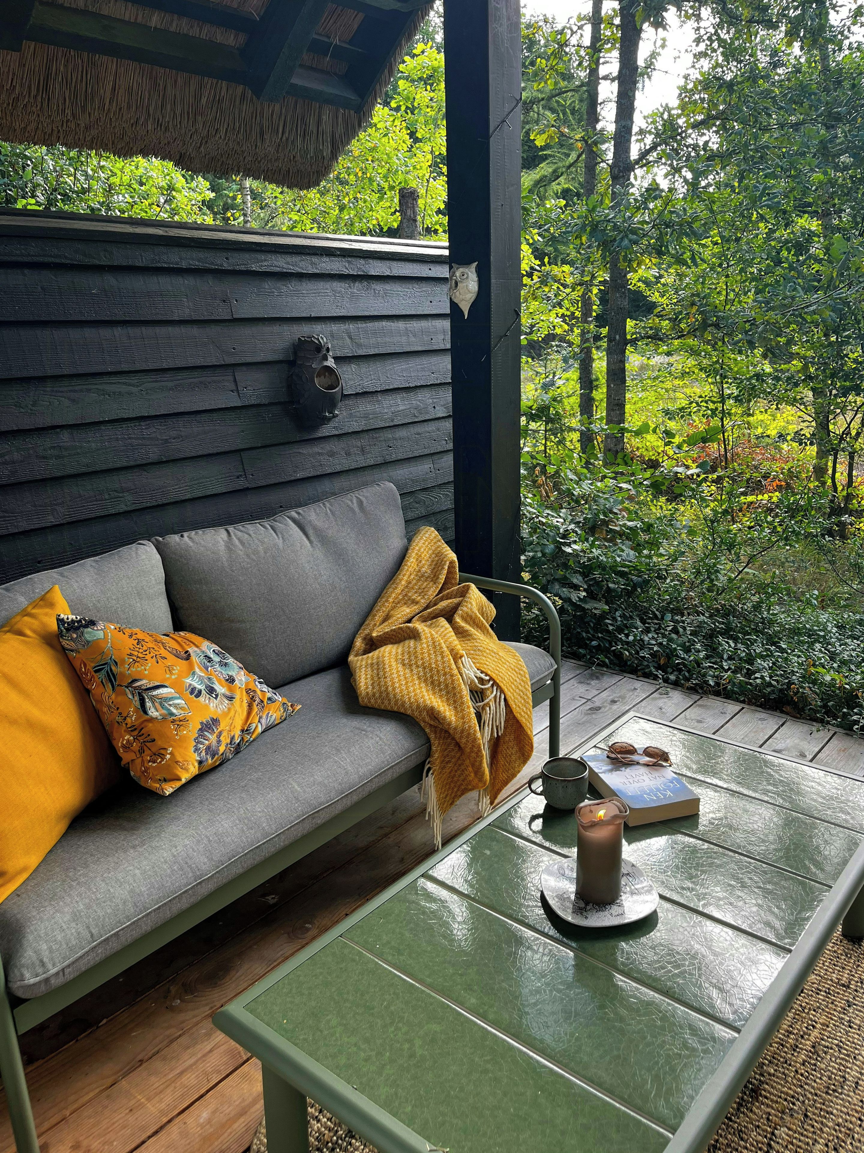 A wooden deck with a grey sofa, a yellow throw blanket, a book, and a mug.