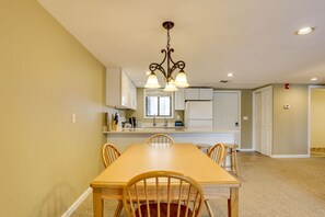A dining area with a wooden table and chairs, a kitchen with white cabinets, and a hanging light fixture.