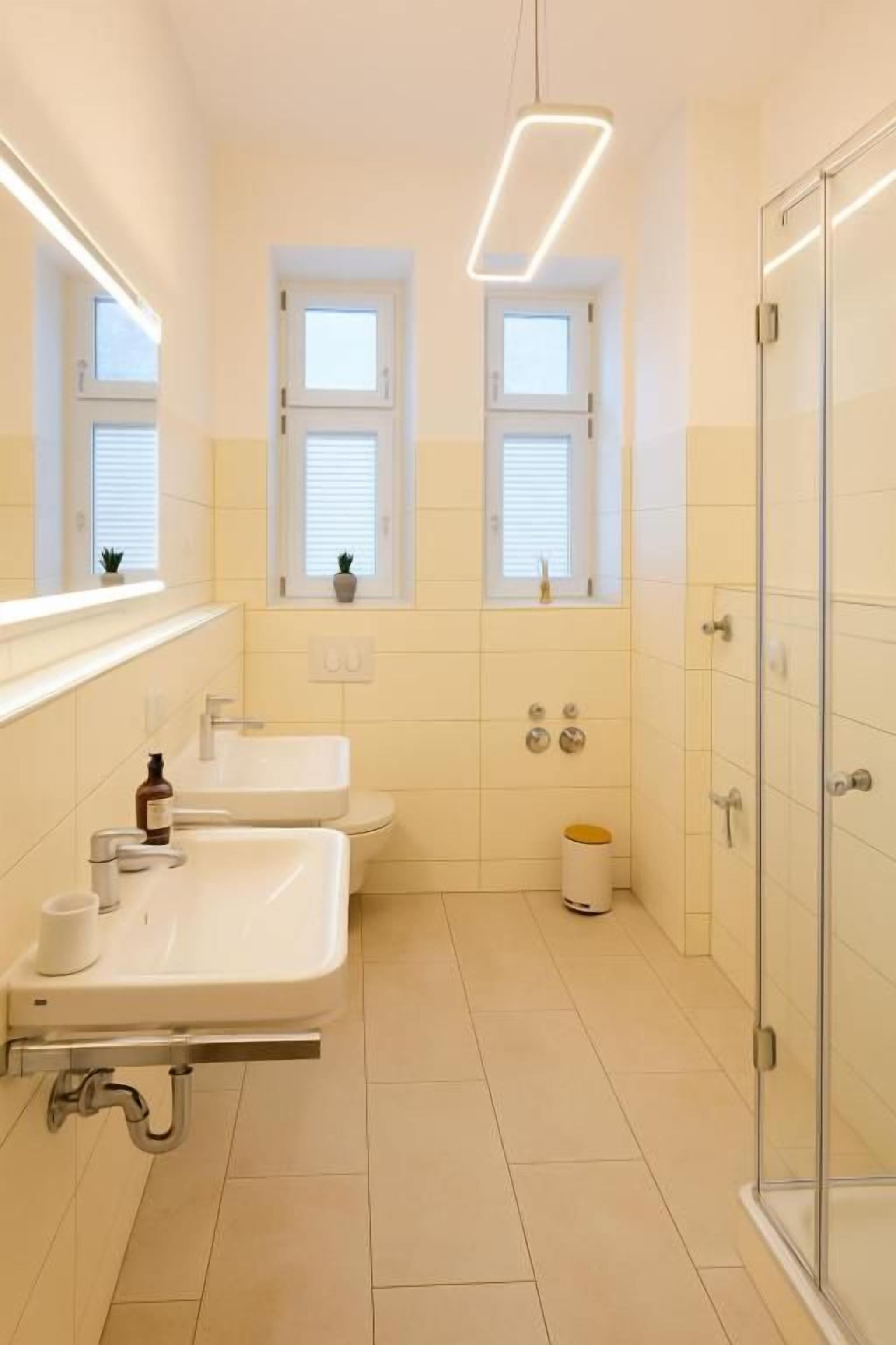 A bathroom with two sinks, a toilet, a glass shower enclosure, and a window with blinds.