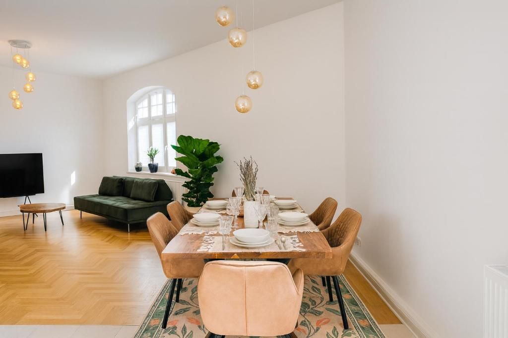 A dining area with a wooden table set for six, surrounded by cushioned chairs. A large plant and a television are visible in the background.