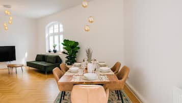 A dining area with a wooden table set for six, surrounded by cushioned chairs. A large plant and a television are visible in the background.