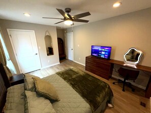 Quarto com cama, escrivaninha com computador, ventilador de teto e televisão.
