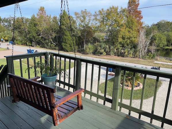 A wooden swing chair on a green deck with a view of a lake and trees.
