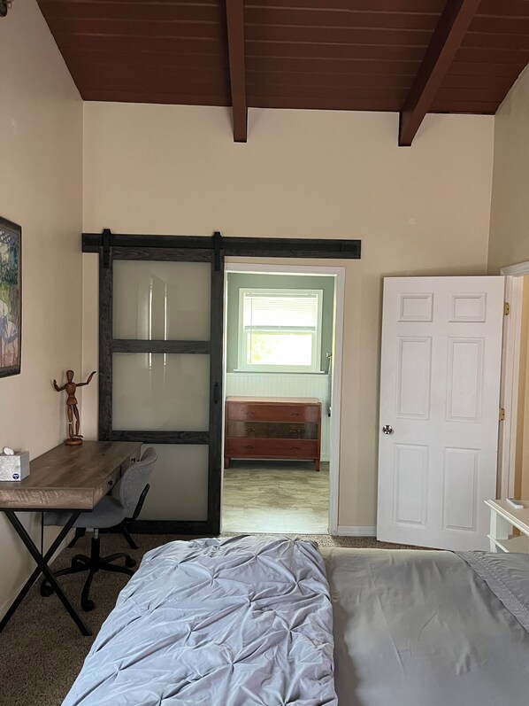 A bedroom with a bed, a desk with a chair, a dresser, and a door leading to another room.
