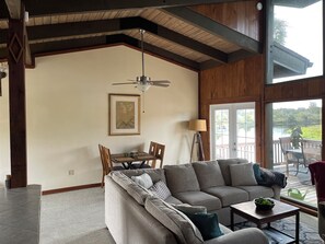 A spacious living room with a gray sectional sofa, a dining table with chairs, a ceiling fan, and a large window offering a view of a lake.