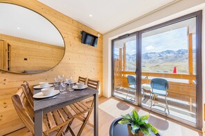 Ein Essbereich mit Tisch für vier Personen, einer Holzwand, einem über dem Tisch montierten Fernseher und einer großen Glasschiebetür, die auf einen Balkon mit Bergblick führt.