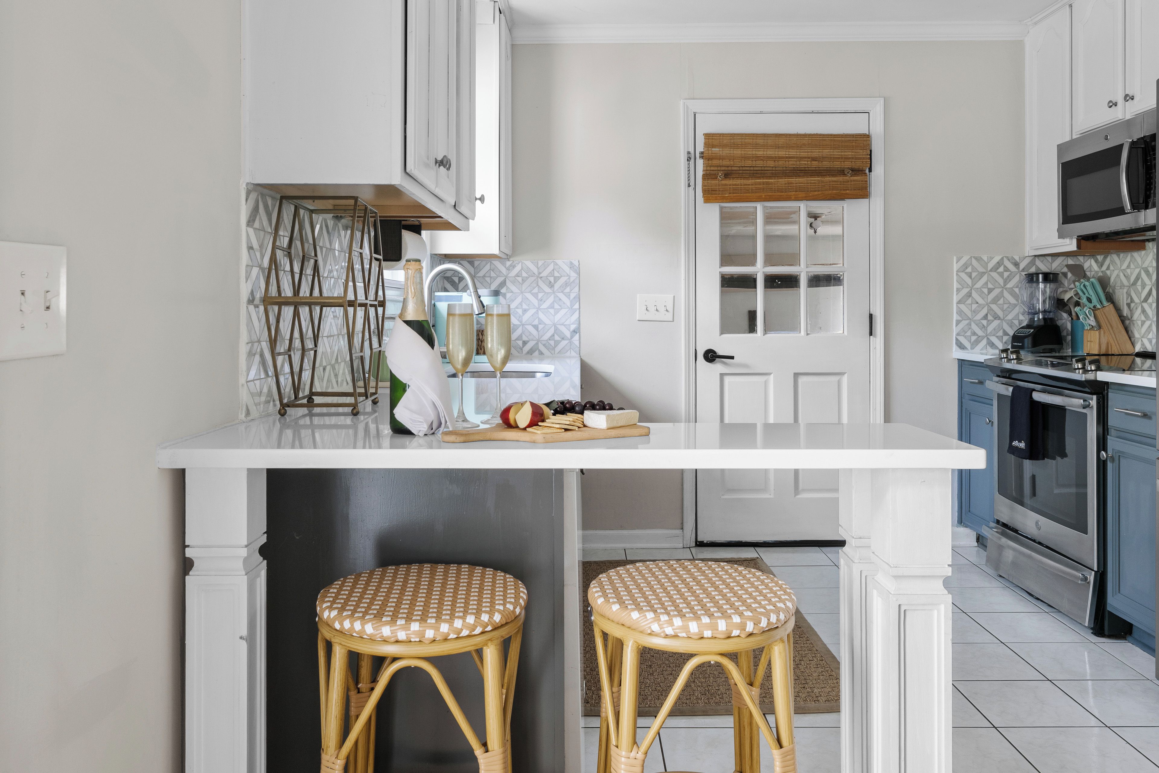 A kitchen with a white countertop, two wicker stools, a door with a window, and a microwave.