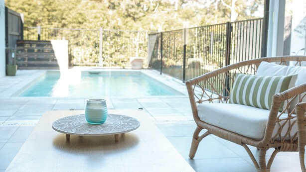Uma área na beira da piscina, com cadeira de vime e mesa, vista desobstruída da piscina e cerca delimitando o espaço.