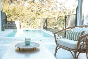 A poolside area with a wicker chair and a table, a clear view of the pool, and a fence enclosing the space.