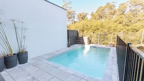 Uma piscina retangular com um deck ladrilhado, cercada por uma cerca baixa, e plantas em vasos à esquerda.