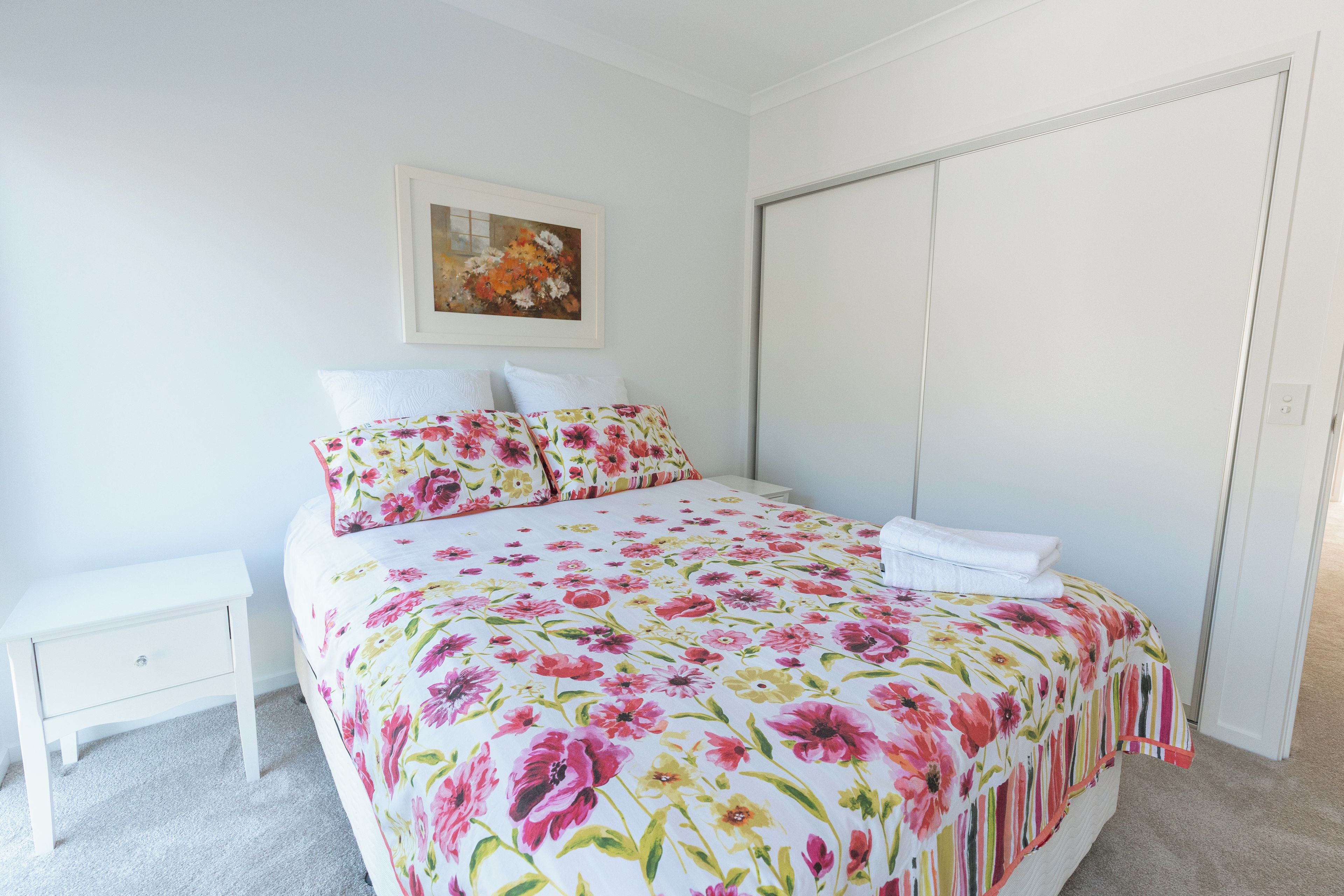 Een slaapkamer met een bed, nachtkastje, schilderij aan de muur en een kledingkast.