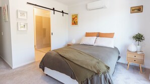 Quarto com cama, mesas de cabeceira, um criado-mudo, ar condicionado fixado na parede, uma porta e um quadro emoldurado.