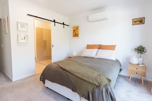 Une chambre à coucher comprenant un lit, des tables de chevet, une commode, un climatiseur fixé au mur, une porte et un tableau encadré accroché au mur.