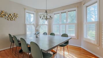 A dining room with a large table, six chairs, a chandelier, and large windows.
