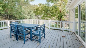 Una terraza de madera con una mesa y sillas, rodeada por una barandilla blanca, con vista a un jardín.