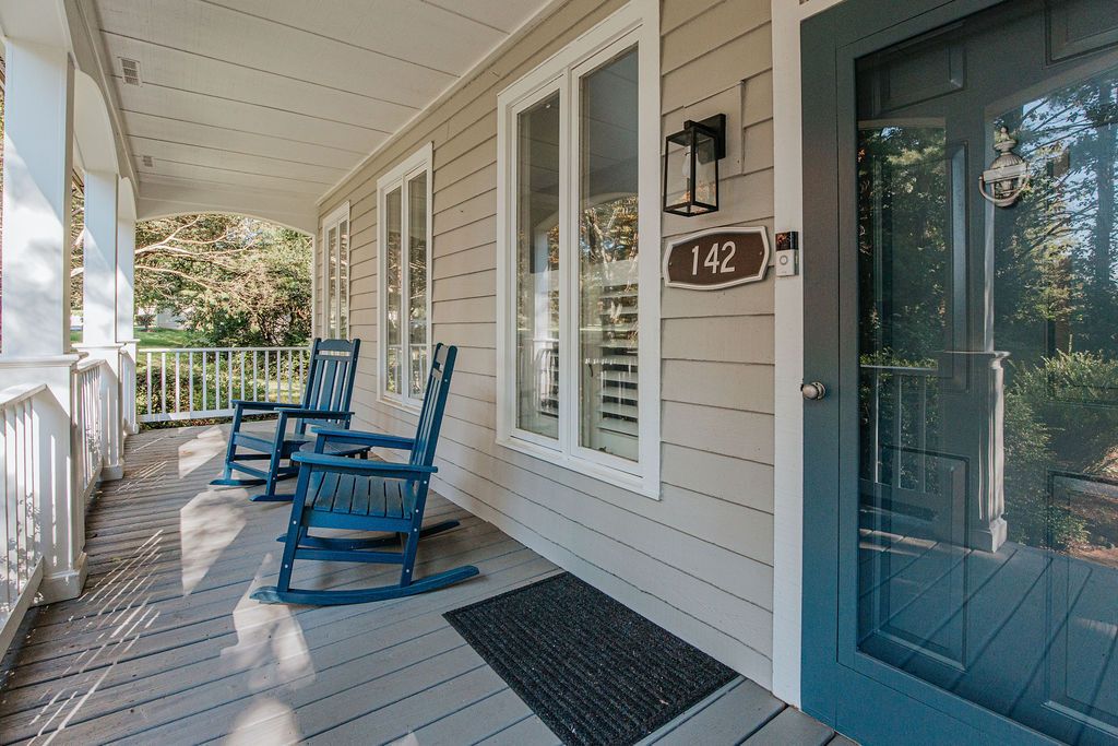 A porch with rocking chairs, a door with a number plate, and a window with a view of trees.
