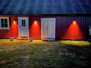 Un edificio rojo con techo de metal corrugado, una puerta blanca y dos ventanas iluminadas.