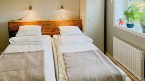 Ett dubbelrum med två sängar, en sänggavel i trä, sänglampor, ett element och ett fönster med växter.