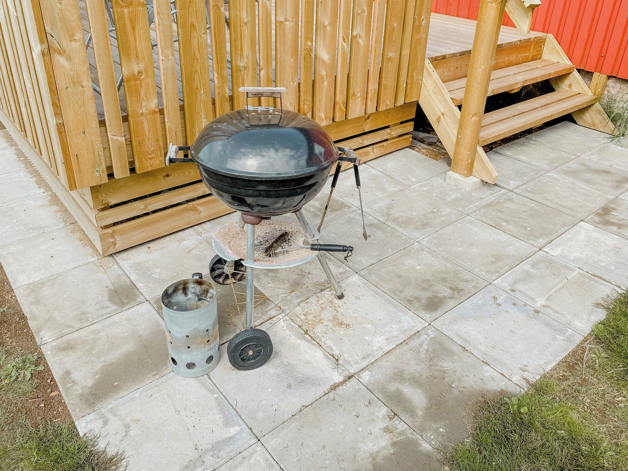 Ein tragbarer Grill mit Metallschornstein und zylindrischem Behälter, auf einer Beton Terrasse mit einem Holzzaun im Hintergrund.