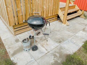 Una parrilla portátil de barbacoa con una chimenea de metal y un contenedor cilíndrico, sobre un patio de concreto con una cerca de madera al fondo.
