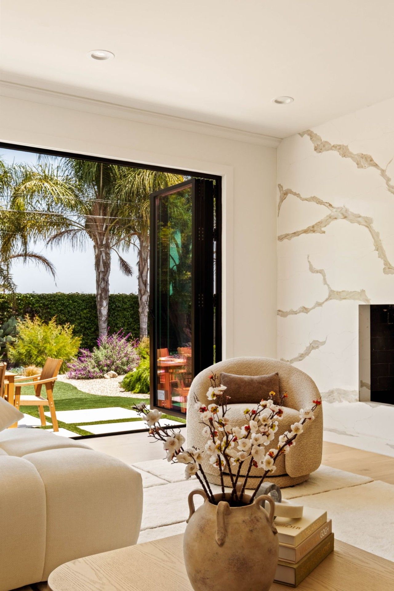 A modern living room with a large glass door leading to a garden, a comfortable sofa, a fireplace, and a decorative vase with flowers.